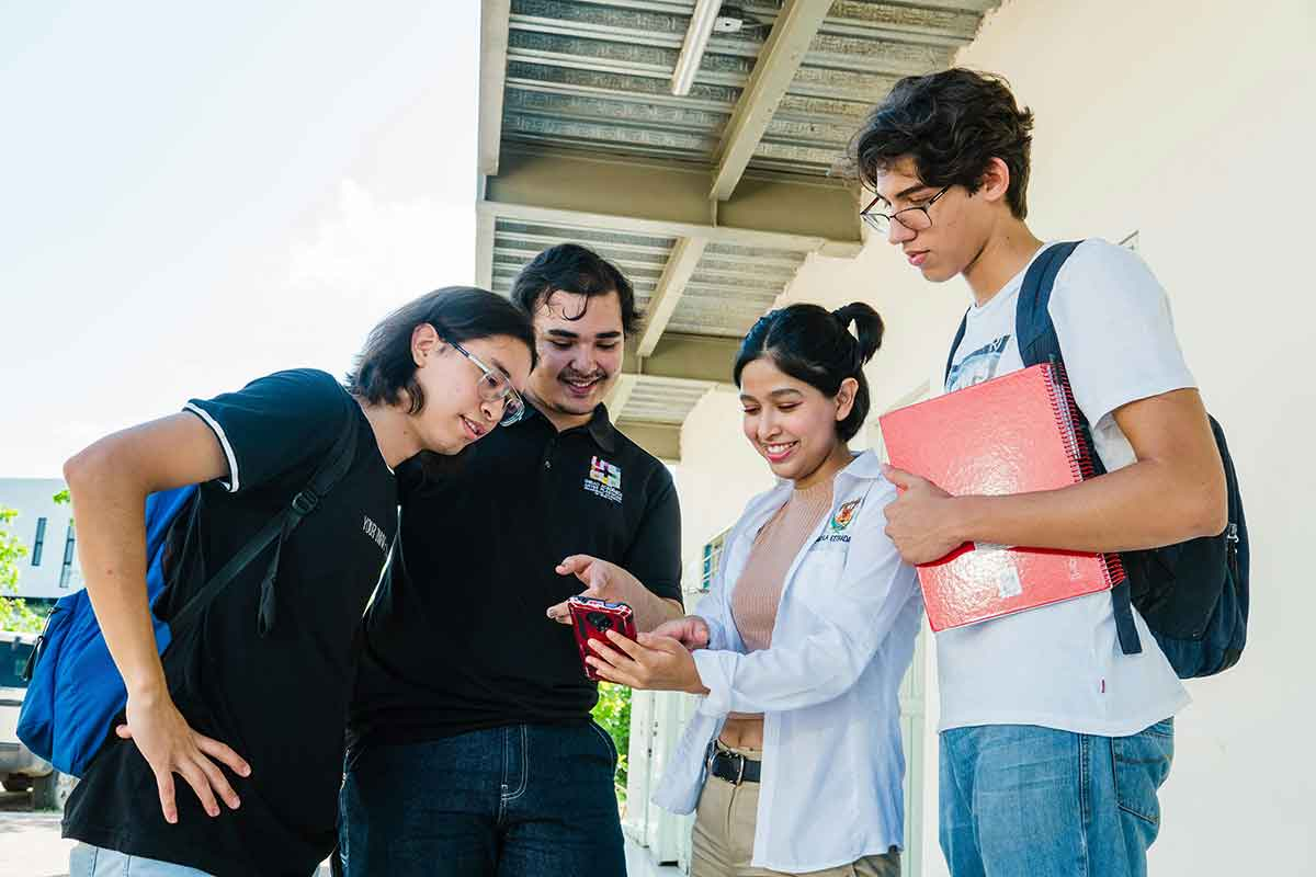 Grupo de estudiantes universitarios sonríe mientras observa un teléfono móvil al aire libre en el campus.
